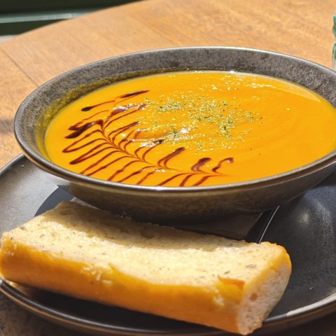 Bowl of orange soup with balsamic drizzle and bread slice on a wooden table.
