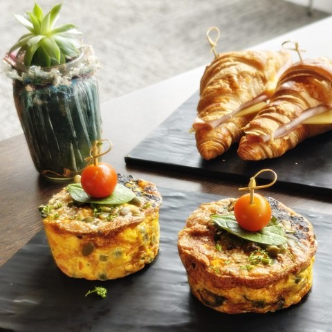 Two quiches topped with cherry tomatoes and two ham croissants on black plates.