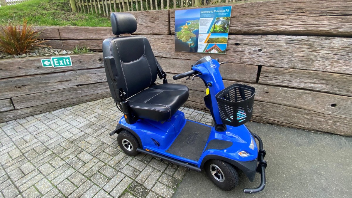 Blue mobility scooter parked on pavement near wooden wall and exit sign.