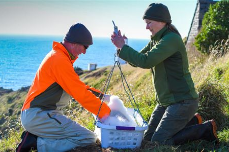 Two people in outdoor gear weighing a basket on a cliff near the sea.